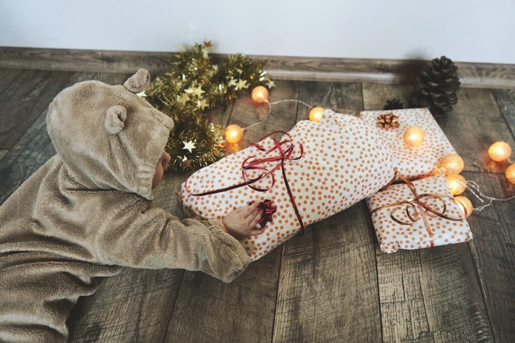 Non-toxic baby holiday image: swaddled infant in bear outfit reaching for an eco-friendly wrapped wooden gift by fairy lights