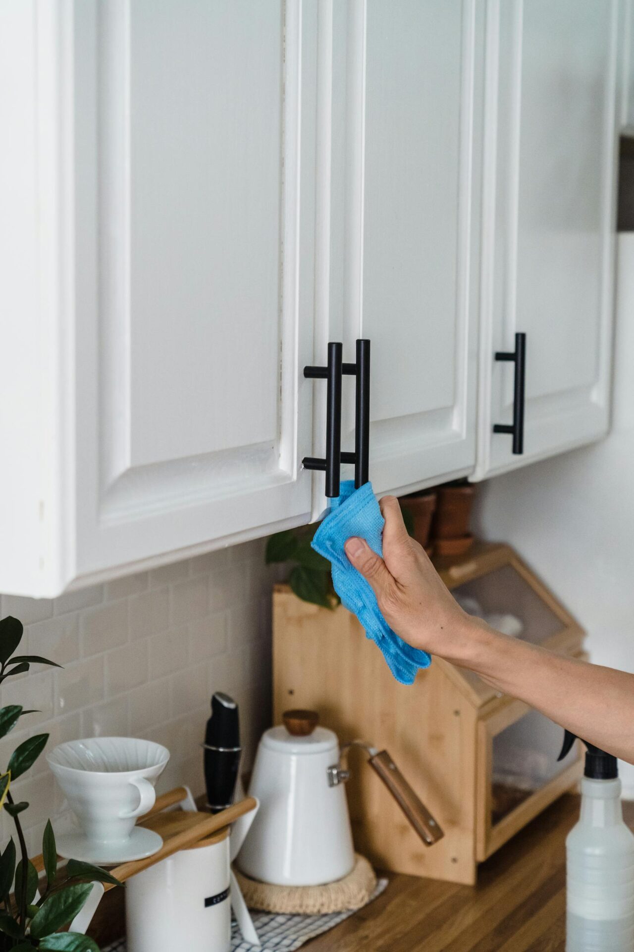 A person using a blue cloth to spring clean white kitchen cabinets with black handles.