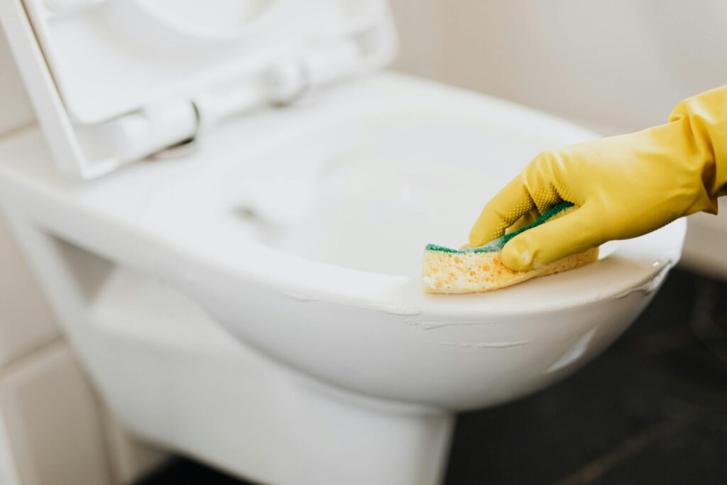 Close-up of a gloved hand using a sponge to clean a toilet, focusing on cleaning products.
