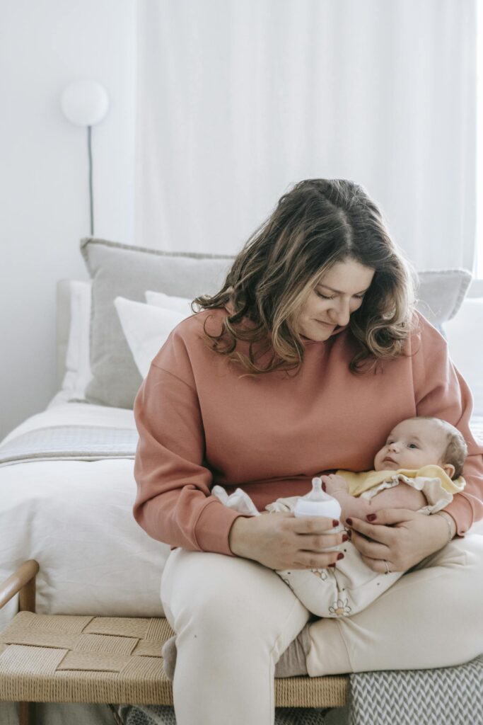 A mother lovingly feeds her baby with a bottle in a cozy, soft-lit bedroom, symbolizing nurturing care.