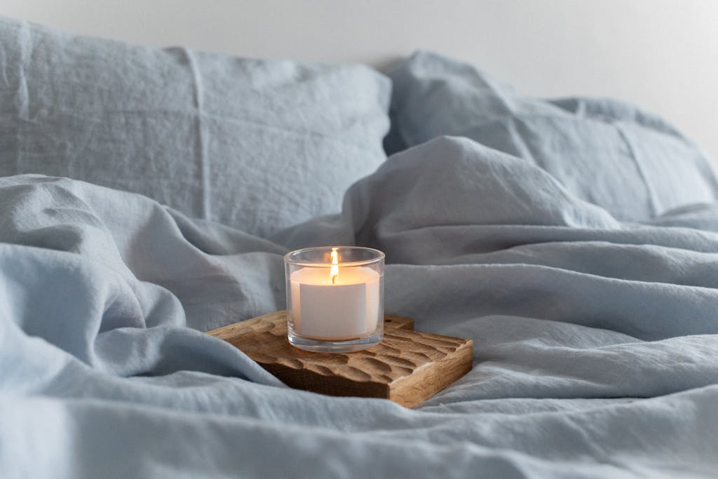 A soothing scene with a lighted candle on a wooden tray amidst soft blue linen bedding.