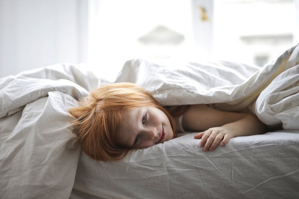 A young girl enjoying a lazy morning, comfortably lying in bed, smiling under a cozy blanket.