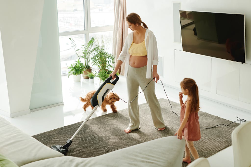 Pregnant mother cleaning carpet with vacuum as daughter and dog play indoors.