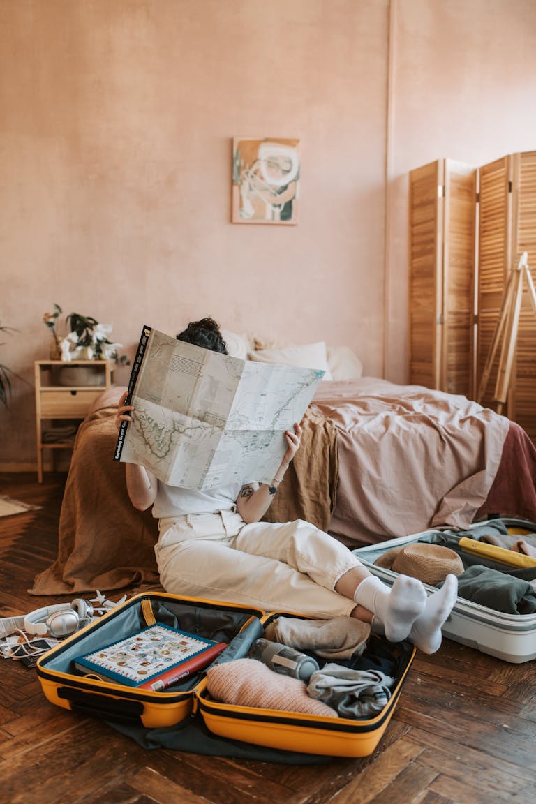 A woman sitting on the floor reading a map, surrounded by non-toxic travel essentials and a packed suitcase in a cozy bedroom.