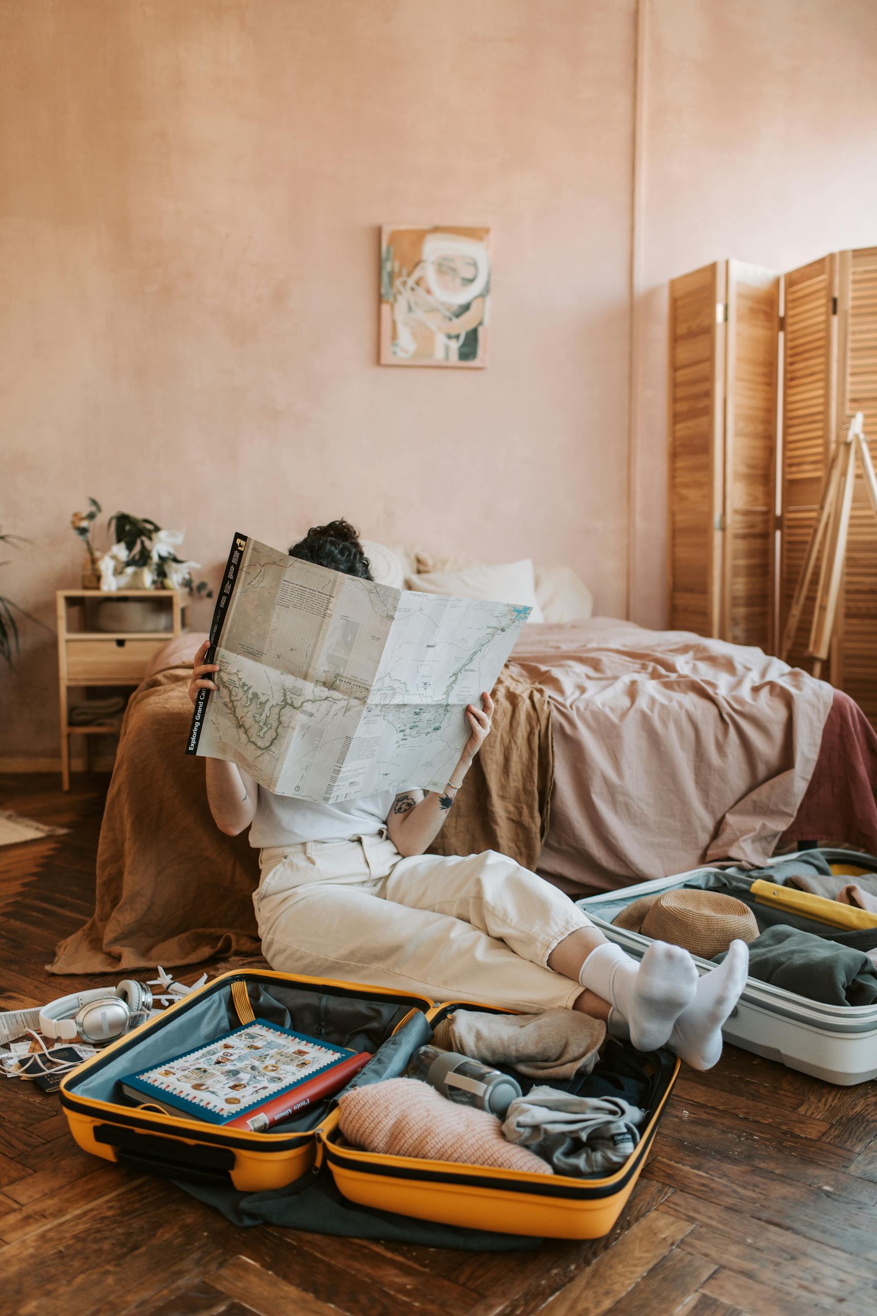 A woman sitting on the floor reading a map, surrounded by non-toxic travel essentials and a packed suitcase in a cozy bedroom.