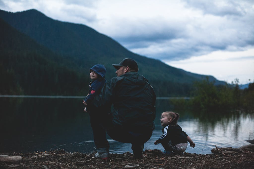Father and children enjoy a serene moment by a lakeside with mountains, protected by non-toxic bug spray.