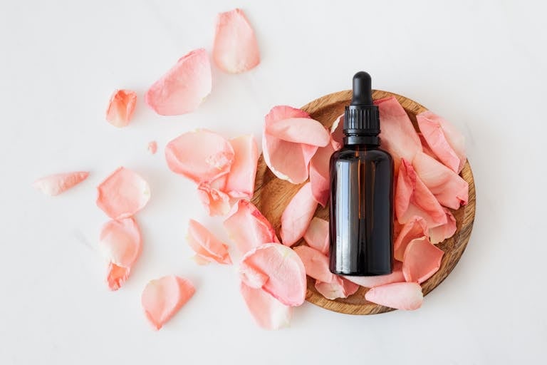 Top view of empty brown bottle for natural skin care product placed on wooden plate with fresh pink rose petals on white background isolated