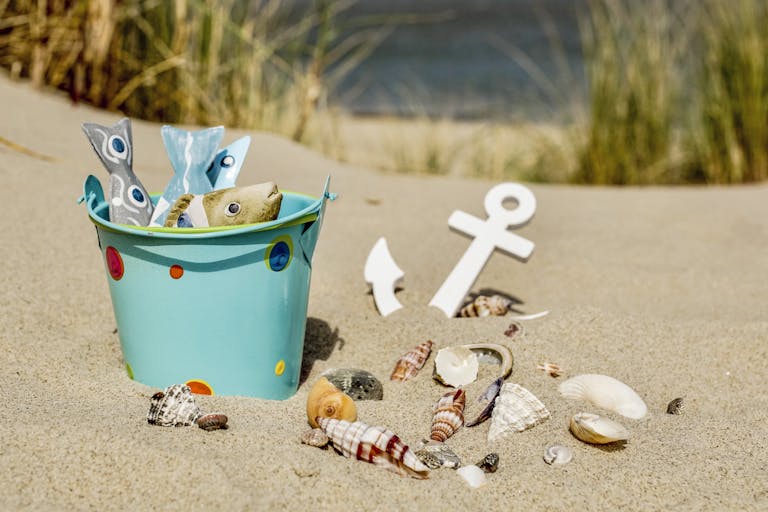 A vibrant beach scene featuring a decorative bucket filled with non-toxic beach gear and seashells on golden sand.