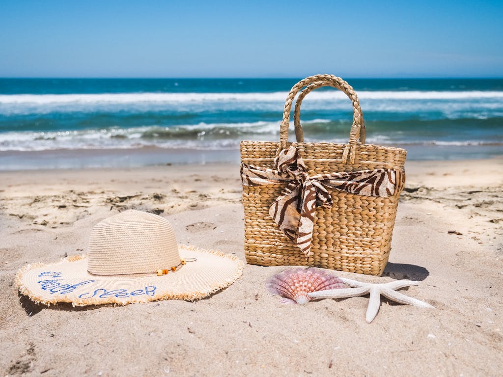 A woven bag and straw hat on sandy beach with ocean waves in the background, perfect for non-toxic summer gear.