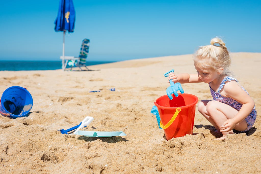 Young girl playing with red bucket and spade on a sunny beach day, enjoying non-toxic summer fun.