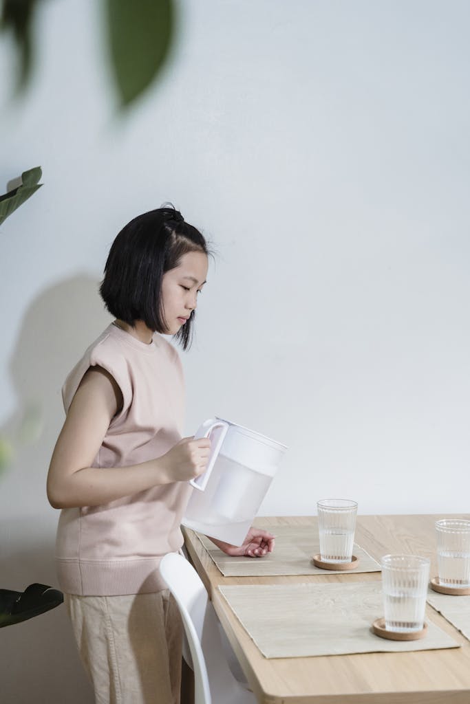A young girl pours water from a pitcher into glasses on a table indoors.