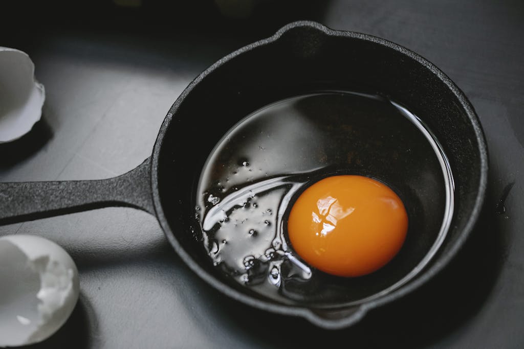 From above of cast iron pan with uncooked egg yolk and white placed on table near scattered shells in kitchen