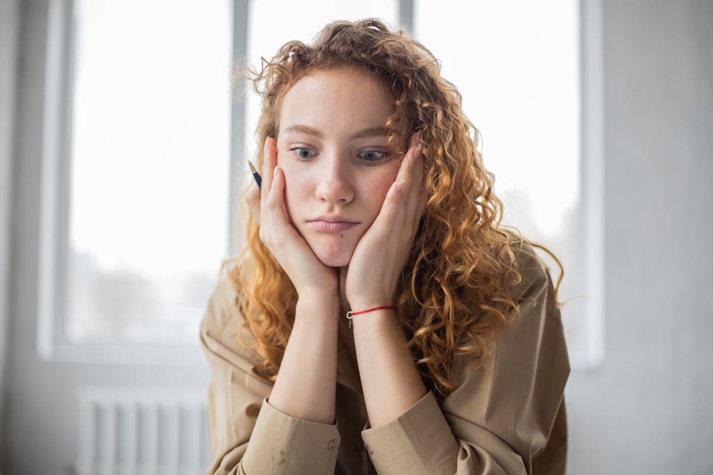 Pensive female student with hands on cheeks looking down while sitting in light room on blurred background while overwhelmed with non-toxic living for beginners.