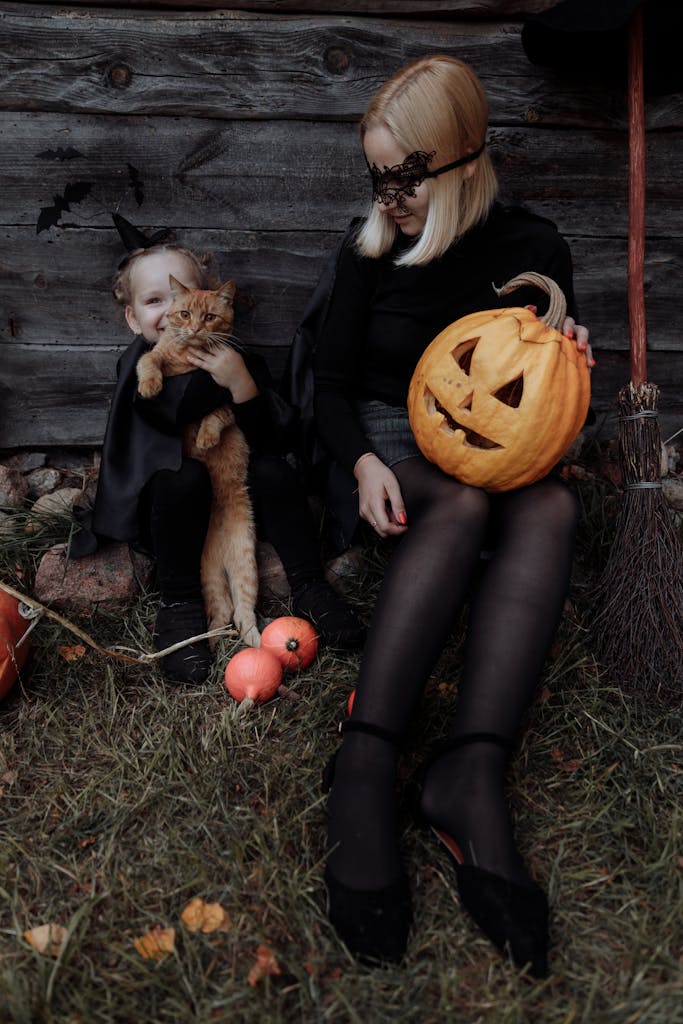 Mom and daughter in witch costumes celebrate Halloween with pumpkins and a cat outdoors.