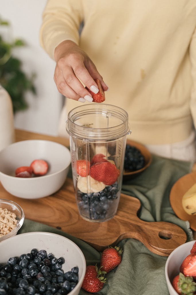 A person putting strawberries into a blender cup with fresh fruit ingredients.