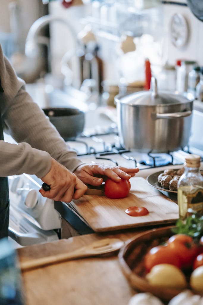 Crop unrecognizable person cutting ripe tomato while preparing lunch against gas stove in house