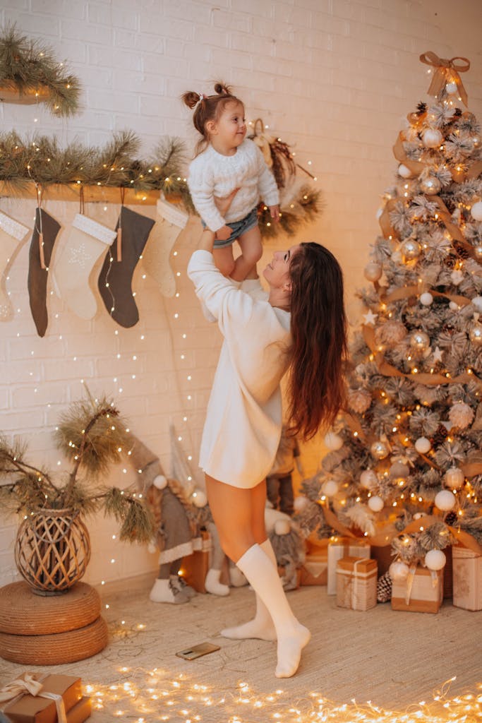 Festive indoor Christmas scene with a mother lifting her child by a decorated tree.