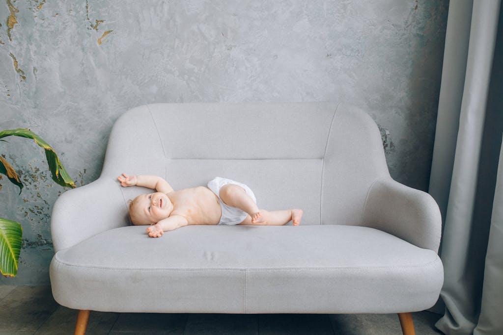 A cute baby in a non-toxic diaper smiling and lying on a stylish gray sofa indoors.