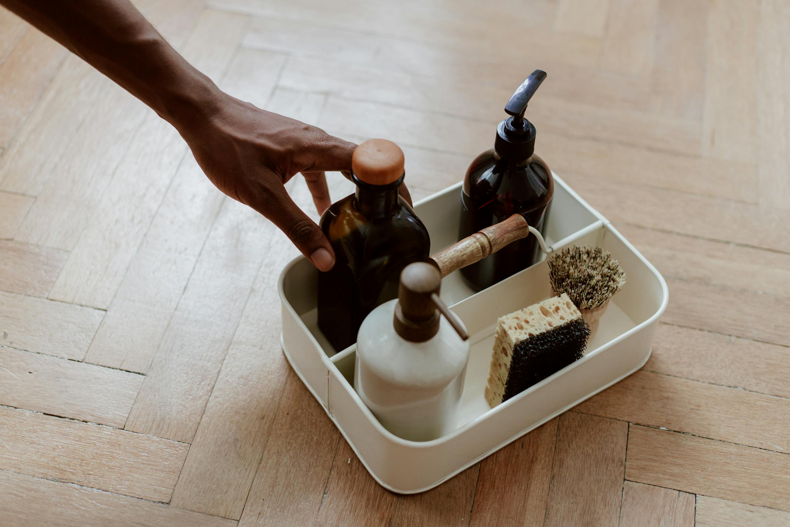 A hand reaching for non-toxic cleaning products with bottles and brushes on a wooden floor.