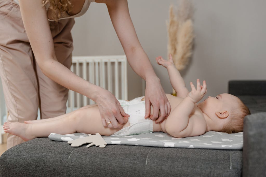 A mother is gently changing her baby's non-toxic diaper on a gray mat in a cozy indoor setting.