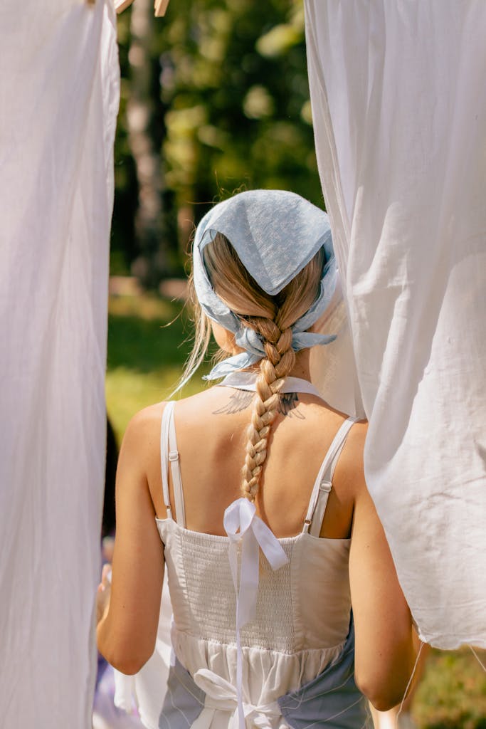 Back view of woman with braided hair and headscarf hanging white laundry outdoors in a sunny rural setting, apron visible