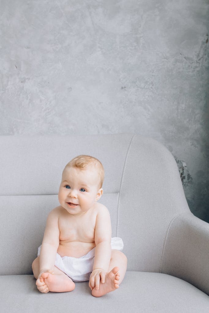Cute baby with short hair in diaper smiling while sitting on a gray sofa indoors.
