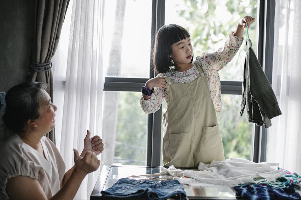 Happy Asian woman showing like gesture while granddaughter organizing clothes on hanger at home.