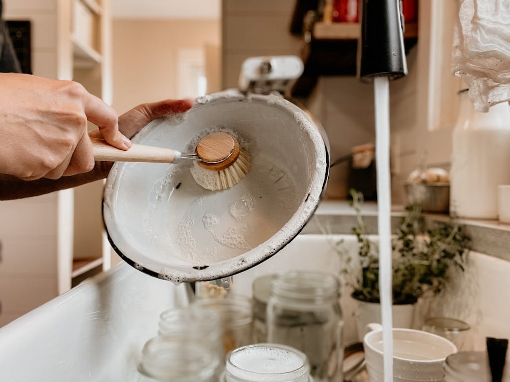 A person hand washing a bowl with a scrubbing brush and non-toxic dish soap in a home kitchen sink. Water is running from the faucet.