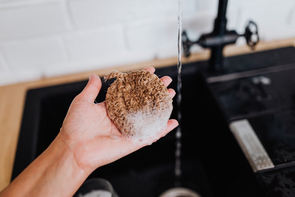 Close-up of a hand holding a natural sponge with non-toxic dish soap under running water at a sink.