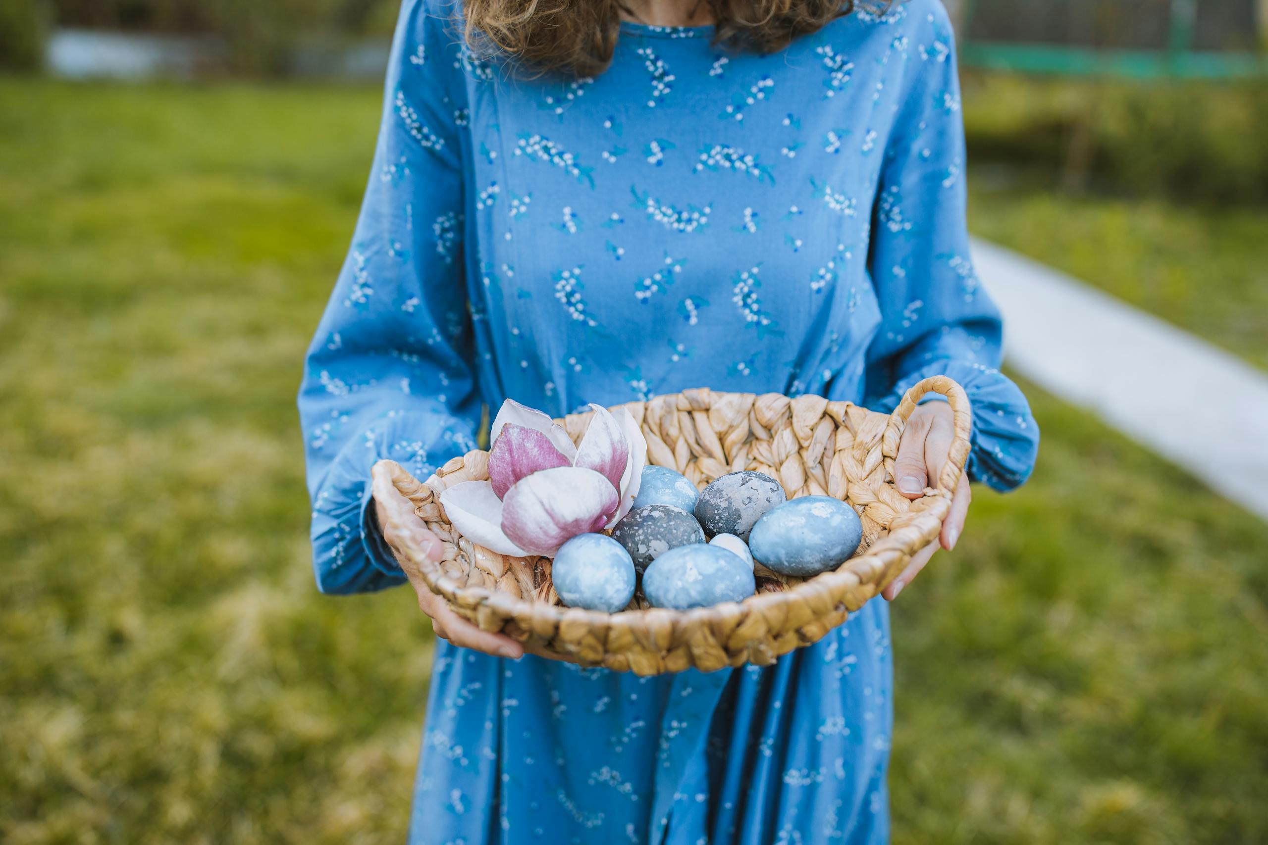 A woman in a blue dress holds a basket of colorful Easter eggs in an outdoor garden setting.