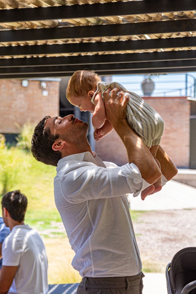 A father joyfully lifts his baby up into the air in an outdoor setting filled with sunlight.