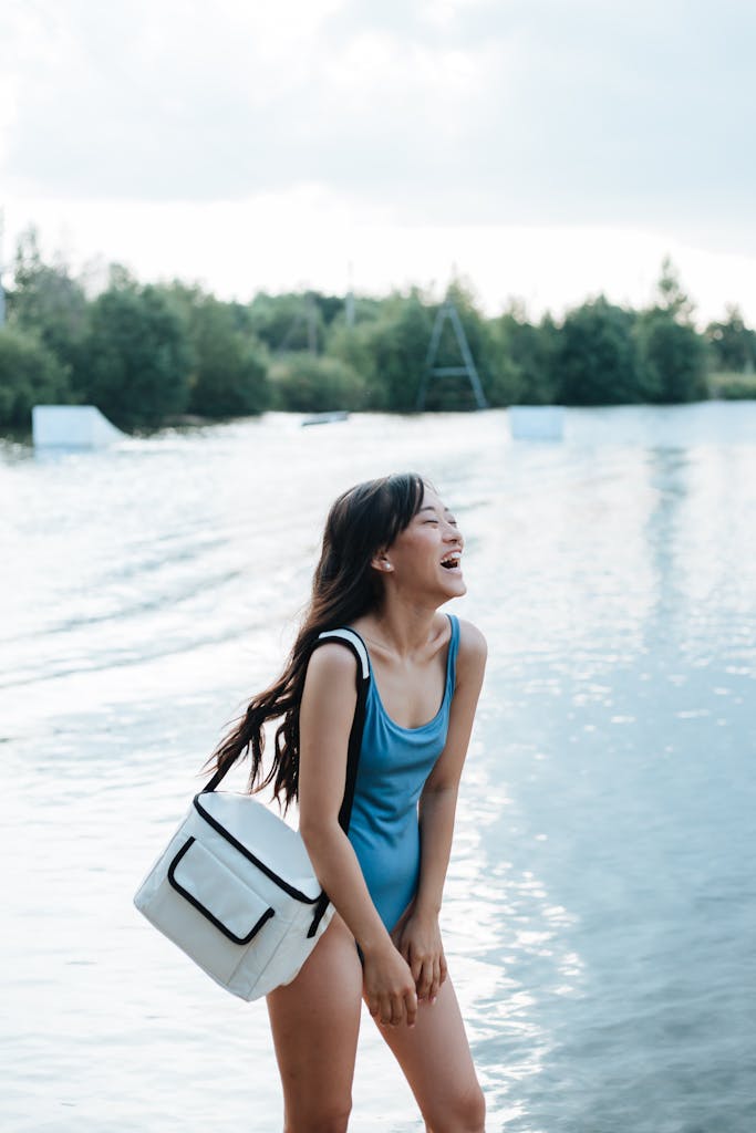 Laughing woman at a lakeside in a blue swimsuit carrying a PFAS-free cooler bag, enjoying a sunny beach day with family gear