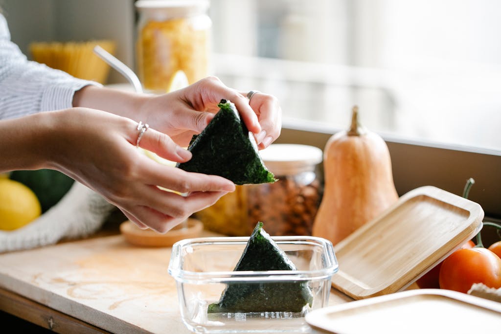 Crop anonymous female cook putting Japanese onigiri wrapped in nori into glass container while standing near counter with assorted food in kitchen on blurred background