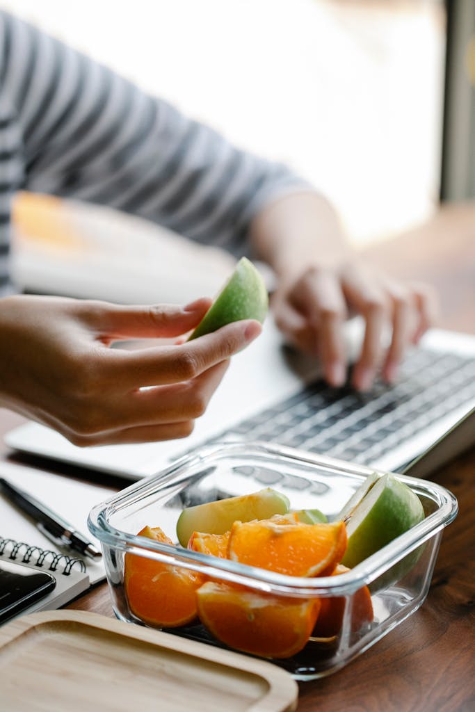 Woman at laptop packing fresh apple and orange slices into a clear non-toxic glass snack container for a healthy work snack