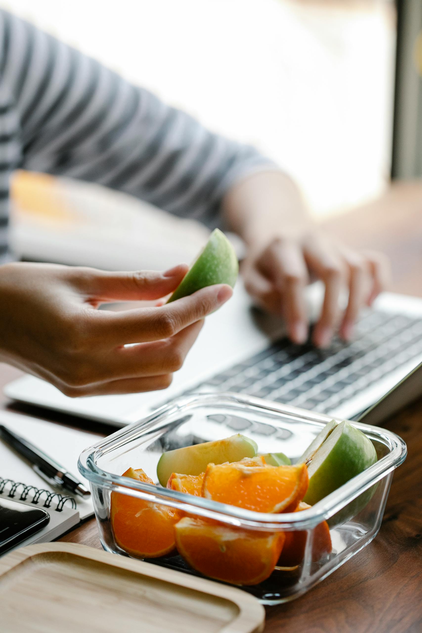 Crop anonymous female taking piece of fresh apple out of plastic container with cut apples and oranges while working on laptop