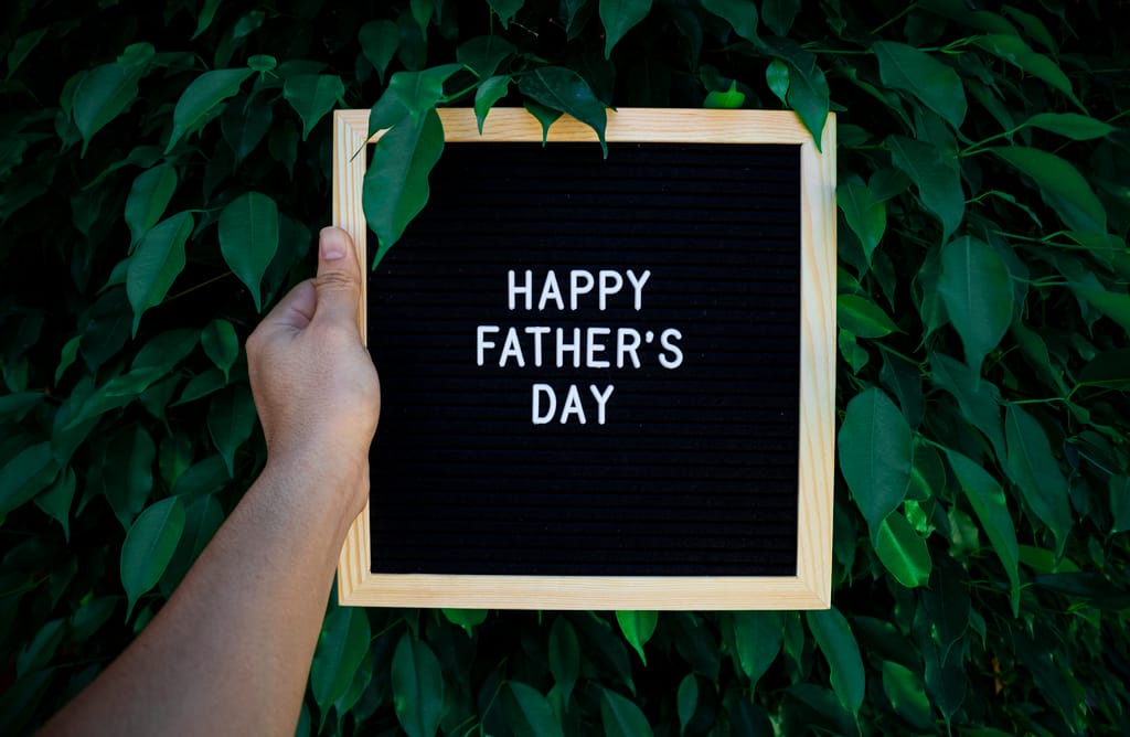 Wooden frame sign with 'Happy Father's Day' text held in front of vibrant green leaves.