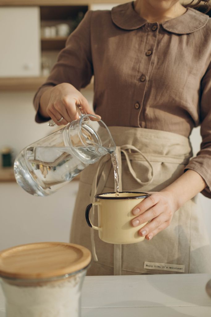 Crop unrecognizable female in cotton casual cloths and beige apron pouring pure water from pitcher into mug while cooking at counter