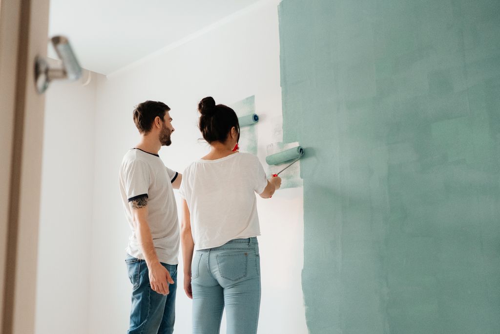 A couple collaborates to paint a wall with a roller indoors, showcasing teamwork.