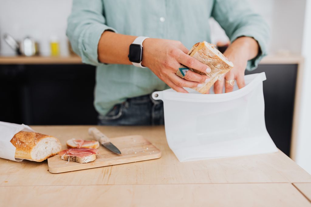 Person placing a sandwich into a white food-grade silicone reusable lunch bag on a kitchen counter showing a PFAS-free option