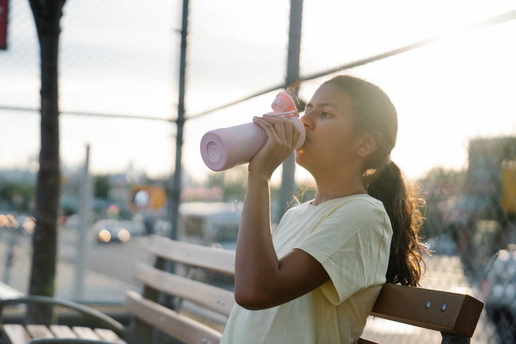 A young girl in a yellow shirt drinks from a bottle on a park bench during sunset outdoors.
