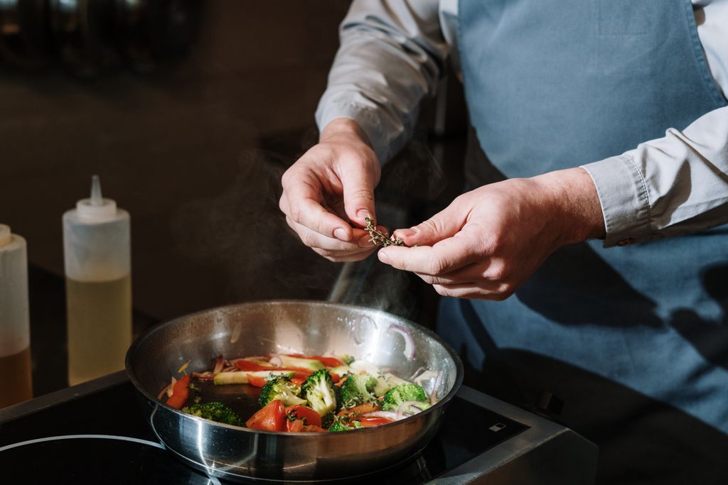Chef seasoning a vegetable stir-fry in a titanium-clad pan, adding fresh herbs over broccoli and tomatoes on a stove