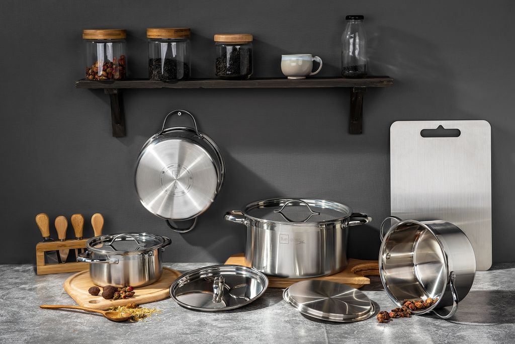Stainless steel cookware set with pots, lids, hanging pan, wooden spoons and glass jars on concrete shelf backdrop