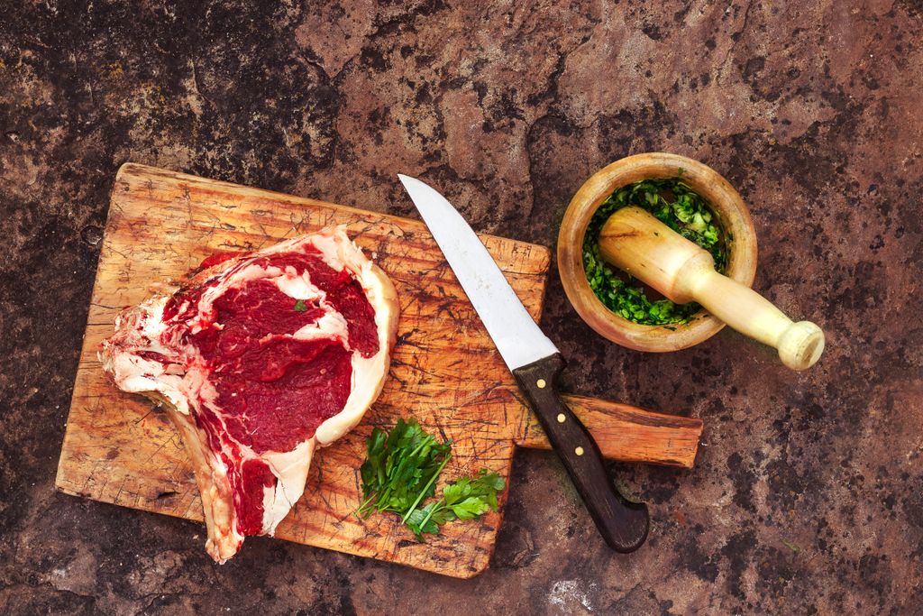 Top view of raw beef steak on a cutting board with herbs and knife on a rustic setting.