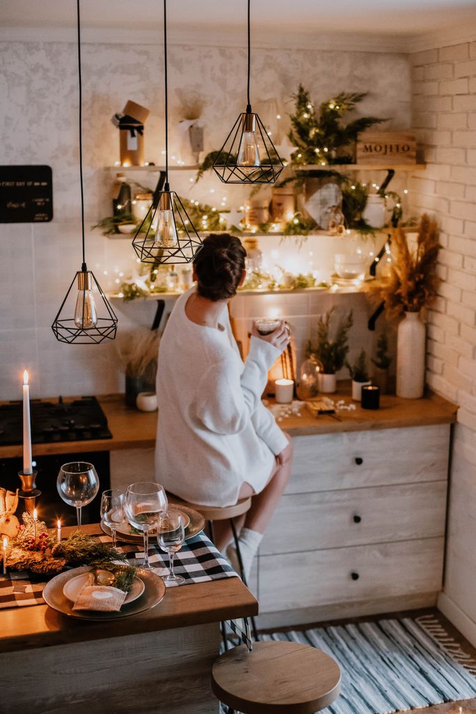 Cozy holiday kitchen scene with a woman holding a mug, warm pendant lights, string lights, candles, and low-tox decor tips