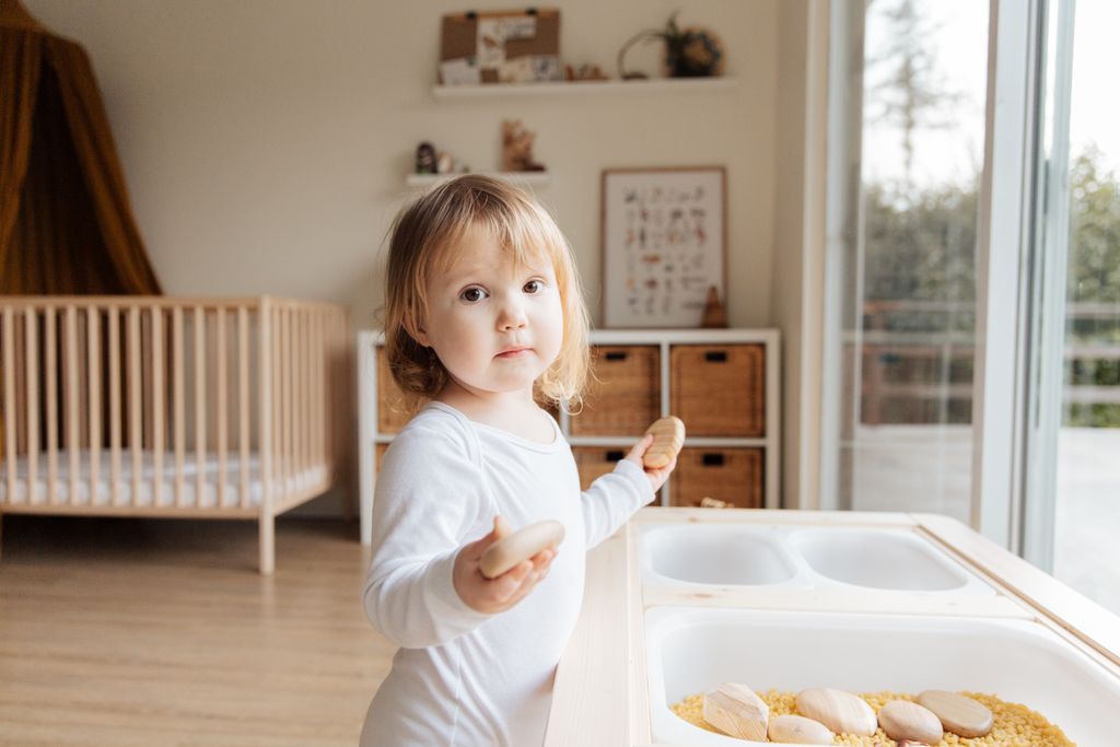 Cute little girl in casual white clothes standing near table with plastic container and playing with stones while looking at camera in modern light room