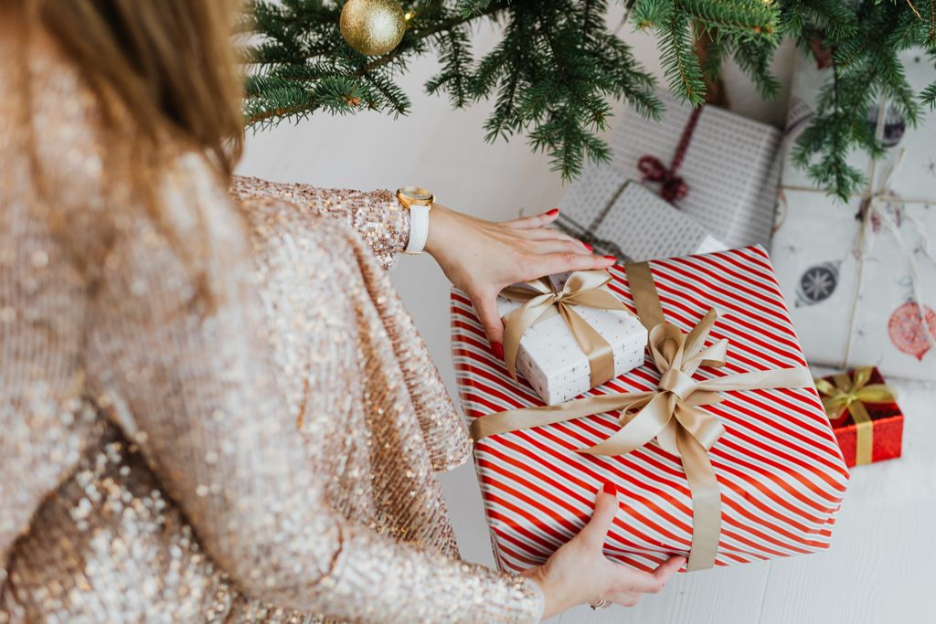 Woman crouching near Christmas tree arranging wrapped gifts at home.